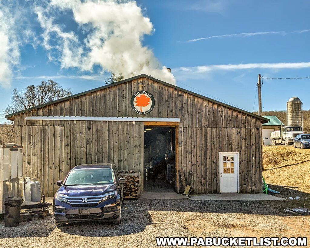 Photo taken during the Somerset County Maple Taste and Tour event showcasing a traditional sugar shack with weathered wooden siding and a distinctive orange maple leaf logo on the front. Billowing white smoke rises from the chimney, indicating maple syrup production inside. A blue Honda car is parked beside neatly stacked firewood, with a white door on the shack's right. The backdrop features a bright blue sky and a farm setting, including a distant silo.