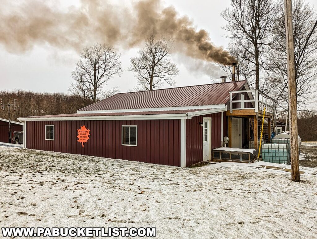 A sugar shack with a metal roof and a maple leaf logo, captured during the Somerset County Maple Taste and Tour event. Dark smoke is emitted from a chimney, suggesting the maple syrup boiling process is active. The shack features a small balcony with stairs leading up to a second door. Surrounding the building is a light dusting of snow, bare trees, and an overcast sky, conveying a chilly winter day in rural Somerset County.