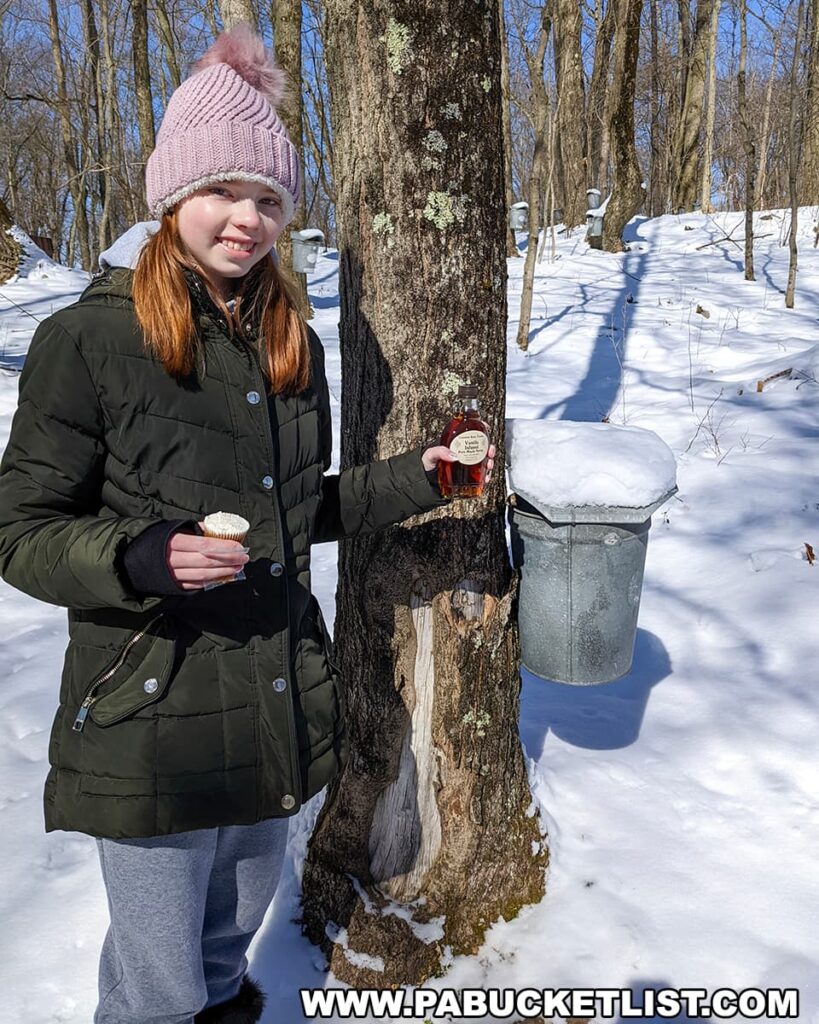 A young girl smiles brightly during the Somerset County Maple Taste and Tour, standing in a snowy forest beside a tapped maple tree. She is dressed warmly in a green winter coat and a pink beanie with a fluffy pom-pom, holding a small bottle of amber maple syrup in one hand and a sample cup in the other. A traditional metal sap bucket is attached to the tree, collecting the dripping sap, ready to be turned into the syrup like the one she proudly displays.