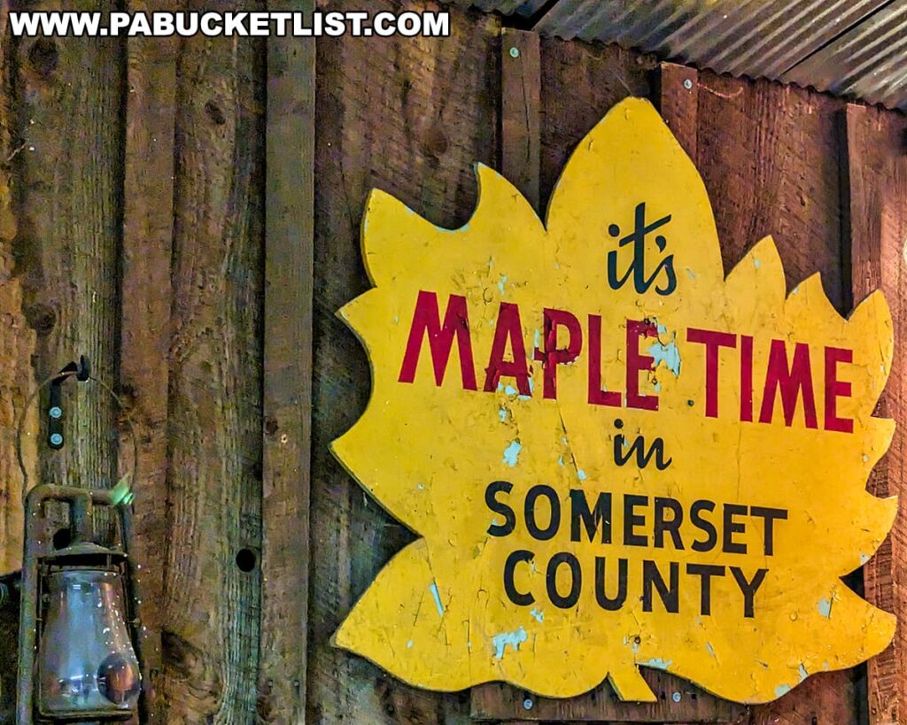 A rustic yellow sign shaped like a maple leaf with the phrase 'it's MAPLE TIME in SOMERSET COUNTY' painted in bold letters, captured during the Somerset County Maple Taste and Tour event. The sign, showing signs of wear with its paint chipping off, is mounted on a weathered wooden wall next to an old-fashioned metal lantern, adding to the charm of the maple syrup celebration atmosphere.