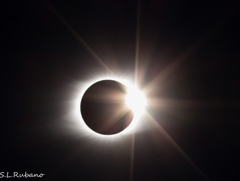 A striking image capturing the moment of a total solar eclipse, with the moon almost completely covering the sun. A radiant burst of sunlight emerges from the side, creating a brilliant diamond ring effect against the dark backdrop of space. The corona of the sun is faintly visible, with rays of light stretching outward.