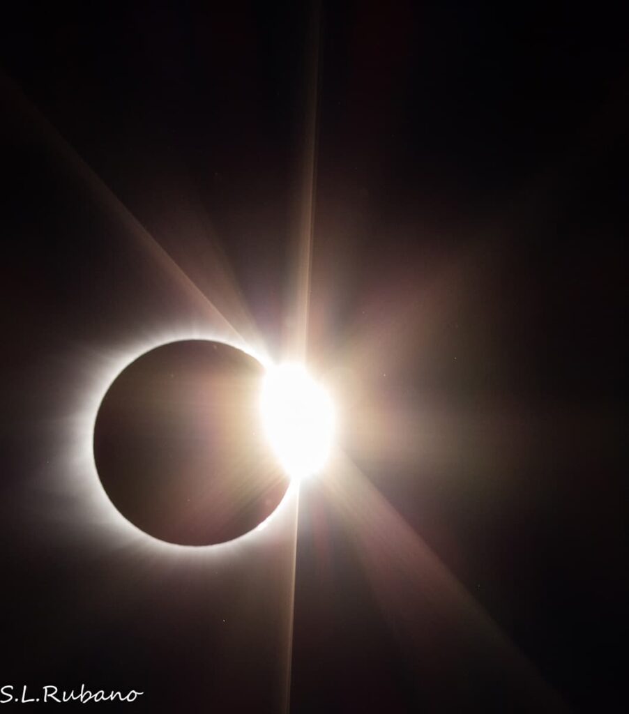 Photograph capturing the dramatic climax of a total solar eclipse, with the sun's radiant corona visible around the silhouette of the moon. A bright flare of light, known as the 'diamond ring effect,' is vividly seen as the sun begins to reemerge. The darkness of space in the background highlights the sun's brilliant halo and the thin stream of light piercing through.