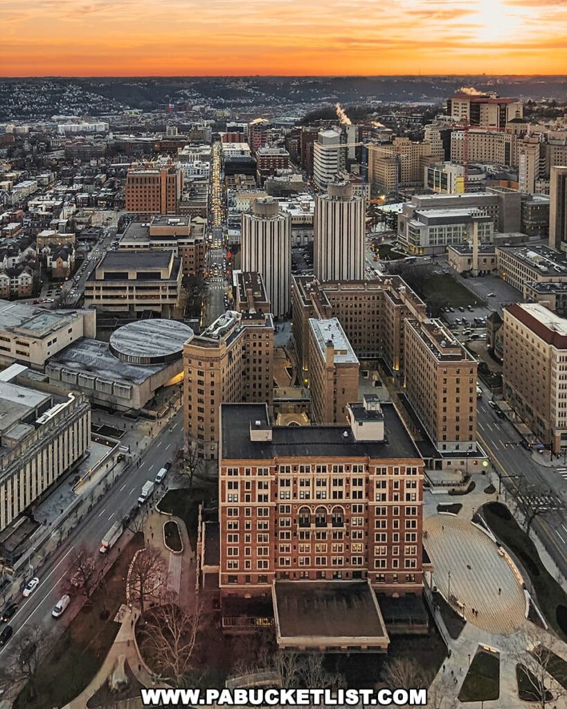 A sunset view over the Oakland neighborhood from the Cathedral of Learning in Pittsburgh, Pennsylvania. The photo captures the dense urban landscape with a mix of modern and traditional buildings, bustling streets, and a radiant orange sky fading into twilight. The city lights begin to punctuate the evening as the day ends, highlighting the vibrancy of the area.