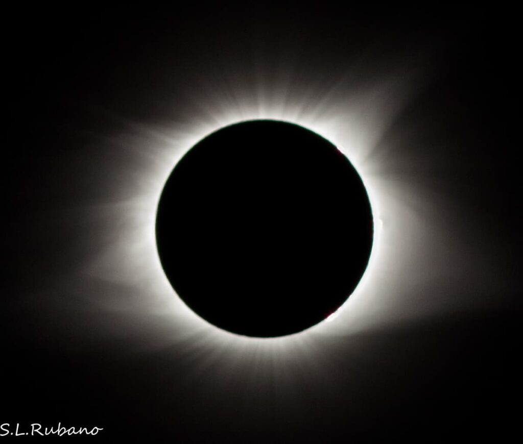 A captivating image of the total solar eclipse, with the moon completely covering the sun, creating a breathtaking corona effect. Rays of sunlight are emanating from the edges of the moon's silhouette, illuminating the surrounding darkness of space. A small red prominence is visible at the edge of the sun's corona.