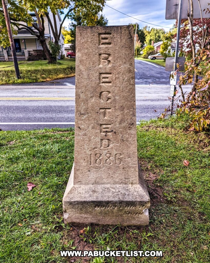 This photo shows the back of a stone marker at the site of the Battle of Gettysburg First Shot Marker along the current Route 30. The marker is inscribed with the word "ERECTED" followed by the date "1886", indicating the year it was established. The stone is standing upright on a concrete base, with a grassy foreground and a paved road running horizontally in the background. Residential houses and trees can be seen in the surrounding area, indicating that the marker is located in a suburban setting. The sky is partly cloudy, suggesting the photo was taken on a day with variable weather.