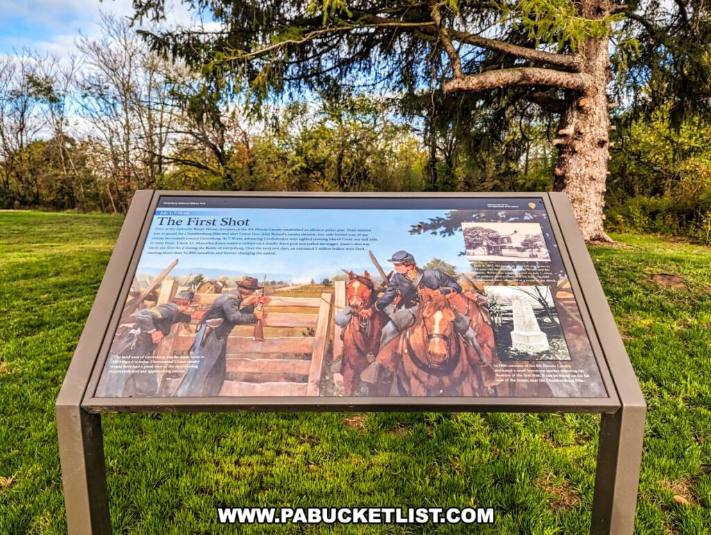 The photo captures an interpretive sign at the Battle of Gettysburg First Shot Marker, set against a natural backdrop with a large tree and a clear sky. The sign details the event of "The First Shot" fired by Lieutenant Marcellus E. Jones of the 8th Illinois Cavalry at the onset of the battle on July 1, 1863. It includes images of the cavalry in action, the Wisler House, and the commemorative marker erected by the veterans of the 8th Illinois Cavalry in 1886. The display is outdoors on a sunny day, providing educational content to visitors about the historical significance of this location along the Chambersburg Pike, surrounded by a peaceful grassy field.