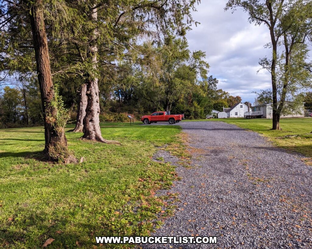 The photo shows a gravel parking area adjacent to the site of the Battle of Gettysburg First Shot Marker. Tall trees with green foliage border the left side of the image, while a red pickup truck is parked on the side of the road, indicating the modern-day use of the area. In the distance, a white house with a covered porch can be seen, possibly the Wisler House, contributing to the serene and historical environment. The setting is peaceful with a clear sky above and hints of residential life, blending the past and present at this significant location along Route 30.