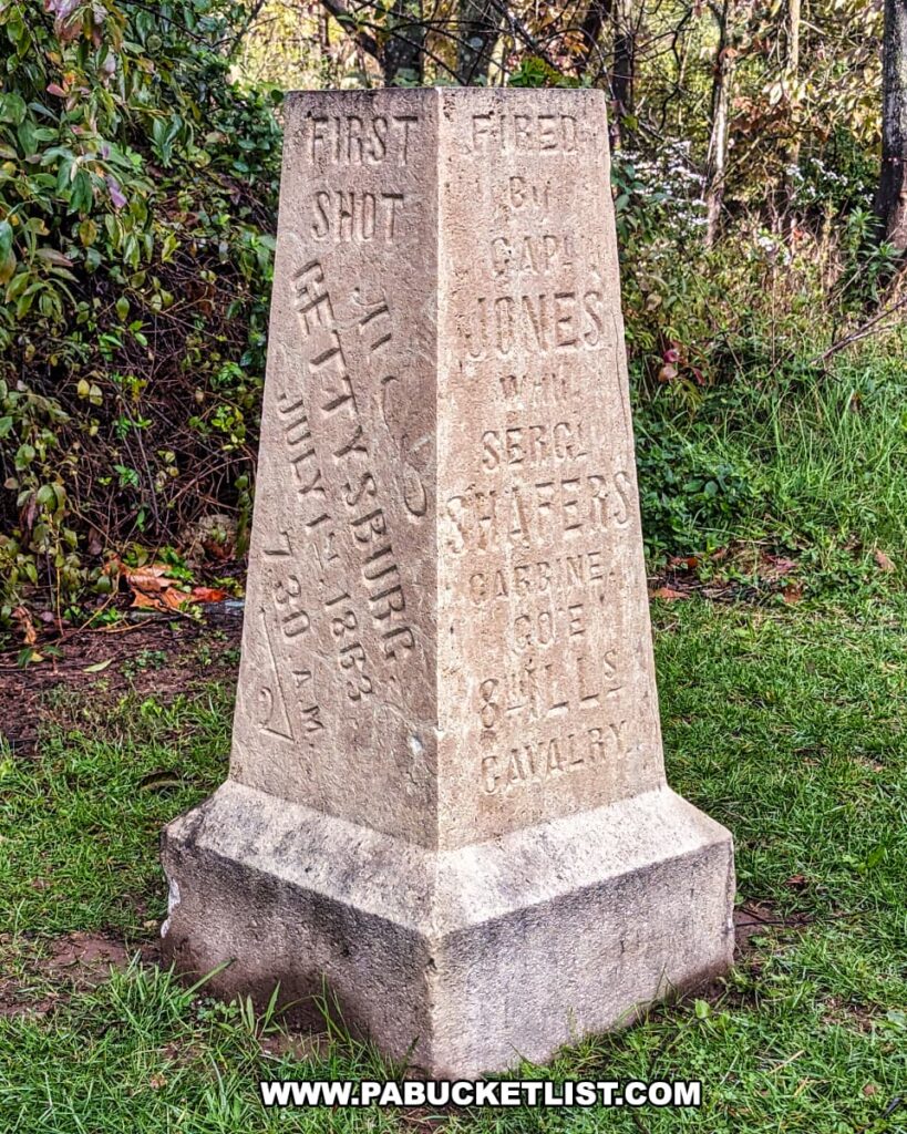 The photo shows the First Shot Marker at the Battle of Gettysburg, standing prominently on a grassy area with a background of lush greenery. Engraved on the limestone monument are the words "FIRST SHOT GETTYSBURG JULY 1, 7:30 A.M. 1863. FIRED BY CAPT. JONES WITH SERGT. SHAFER CARBINE CO. E 8 ILLS. CAVALRY," commemorating the moment Lieutenant Marcellus E. Jones of the 8th Illinois Cavalry fired the first shot of the battle. The stone stands as a solemn reminder of the historical event, etched with the names of those involved, preserving their legacy on the hallowed grounds along Chambersburg Pike.