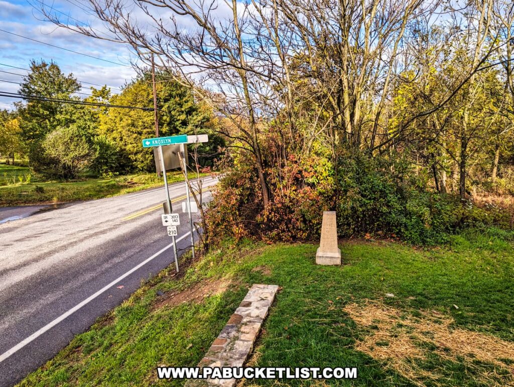 The photo shows a tranquil roadside view near the Battle of Gettysburg First Shot Marker along Route 30. A street sign reads "Knoxlyn Rd," pointing down a road that intersects with the main route. A modest stone marker stands in the foreground amidst green grass and autumnal foliage, marking the significant location where Lieutenant Marcellus E. Jones is said to have fired the first shot of the battle. The overcast sky suggests a quiet, reflective day at this historic site.