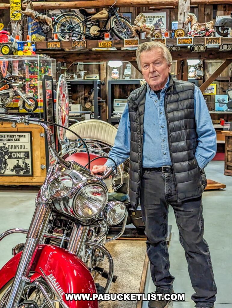 Bill Morris stands confidently in front of a vintage motorcycle at Bill's Old Bike Barn museum in Bloomsburg, Pennsylvania. He's dressed casually in a blue puffer vest over a light shirt, black belt, dark jeans, and sports neatly combed hair. The background reveals an eclectic collection of memorabilia, including mounted bicycles, license plates, toy cars, and framed pictures, showcasing the rich history and ambiance of the museum. The array of collectibles creates a vibrant tapestry that reflects a passion for Americana and the golden era of motorcycles.