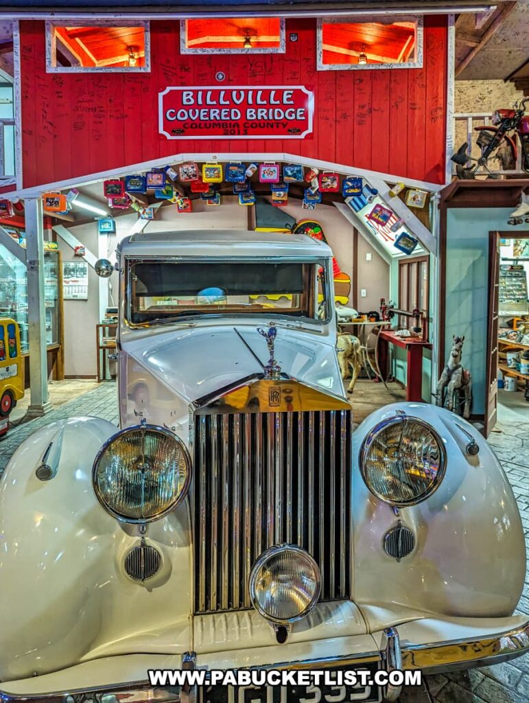 A pristine vintage Rolls-Royce car with a prominent grille and classic round headlamps is on display at Bill's Old Bike Barn museum in Bloomsburg, Pennsylvania. Above it hangs a red sign that reads "BILLVILLE COVERED BRIDGE COLUMBIA COUNTY 2013" flanked by small, lit windows. The space is crammed with nostalgic items, including colorful license plates lining the wooden beam, vintage tin lunch boxes, and assorted memorabilia. A bicycle is perched high on a ledge, contributing to the rich visual history evident throughout the museum. The setting offers a charming look into past eras, inviting visitors into a world where every artifact tells a story.