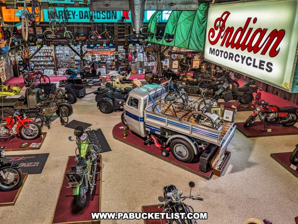 An elevated view of the Military Room at Bill's Old Bike Barn museum in Bloomsburg, Pennsylvania, showcasing a vast collection of vintage motorcycles, automobiles, and military memorabilia. Dominating the scene are prominent signs for Harley-Davidson and Indian Motorcycles, with an array of classic bikes, including some with sidecars and one vivid green motorcycle, lined up below. Vintage bicycles are suspended from the ceiling, and there is an assortment of old wagons, a small trailer, and various military equipment. The collection is densely displayed, offering a treasure trove of historic and cultural artifacts that celebrate motor history and Americana.