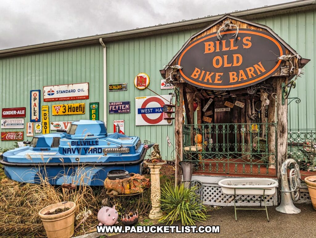 The entrance to Bill's Old Bike Barn museum in Bloomsburg, Pennsylvania, features an eclectic mix of vintage and rustic elements. A prominent oval sign with "Bill's Old Bike Barn" welcomes visitors, set against a weathered wood frame flanked by antique lanterns. Below, a whimsical array of items includes a small blue boat labeled "Billville Navy Yard," a porcelain bathtub, various pots, a piggy bank, and an old musical instrument. The wall behind this collection is adorned with classic gas station and beverage signs, contributing to the nostalgic charm of this unique roadside attraction.