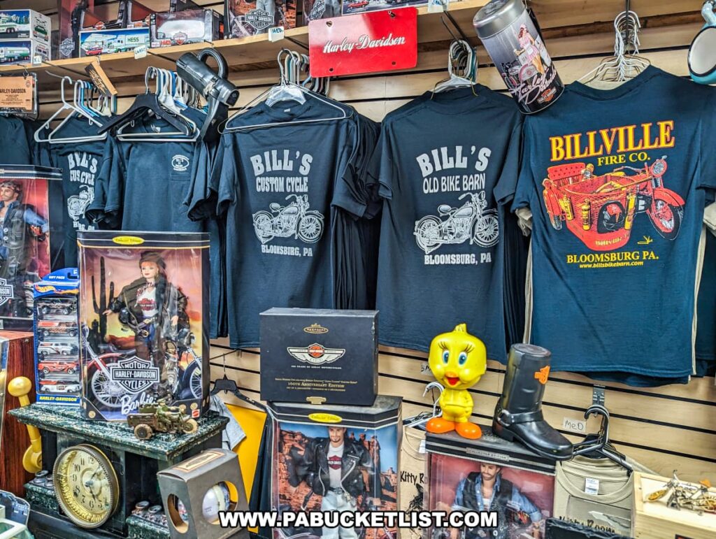 The gift shop at Bill's Old Bike Barn museum in Bloomsburg, Pennsylvania, displays a variety of memorabilia and souvenirs. There are several t-shirts hanging, each with a different design related to the museum, such as "Bill's Custom Cycle" and "Billville Fire Co." Also featured are collector's items including a Harley-Davidson themed Barbie doll in its box, a retro style lunch box hanging above, and various model cars and motorcycles on the shelves. A quirky Tweety Bird figurine and a miniature boot add to the shop's eclectic charm. This area is a treasure trove for visitors looking to take a piece of motorcycle history home with them.
