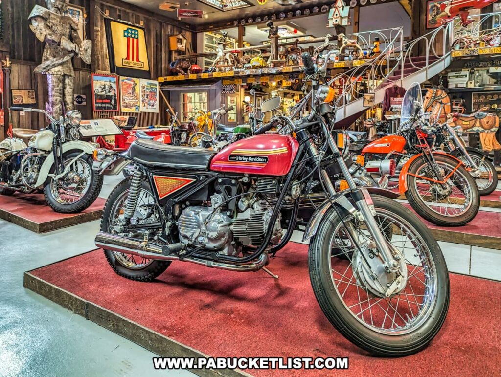 A red vintage Harley-Davidson motorcycle is prominently featured at Bill's Old Bike Barn museum in Bloomsburg, Pennsylvania, part of a larger display of classic bikes. This well-preserved motorcycle is showcased on a red carpet platform, highlighting its sleek lines and chrome details. The backdrop is filled with an array of Americana memorabilia, including mounted bicycles, Americana flags, and varied collectibles on wooden shelves that capture the spirit of the American road and its rich motorcycling history.
