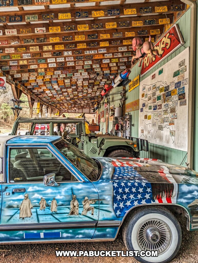 A creatively decorated car at Bill's Old Bike Barn museum in Bloomsburg, Pennsylvania, features a striking mural of the American flag blending into a scene with the Statue of Liberty. Above the car, a collection of colorful vintage license plates from various states forms a canopy. A map of the United States with corresponding state license plates and a sign for "Tony's Bikes" contribute to the diverse and historical backdrop. Military vehicles are also visible, enhancing the rich, patriotic theme of the display.