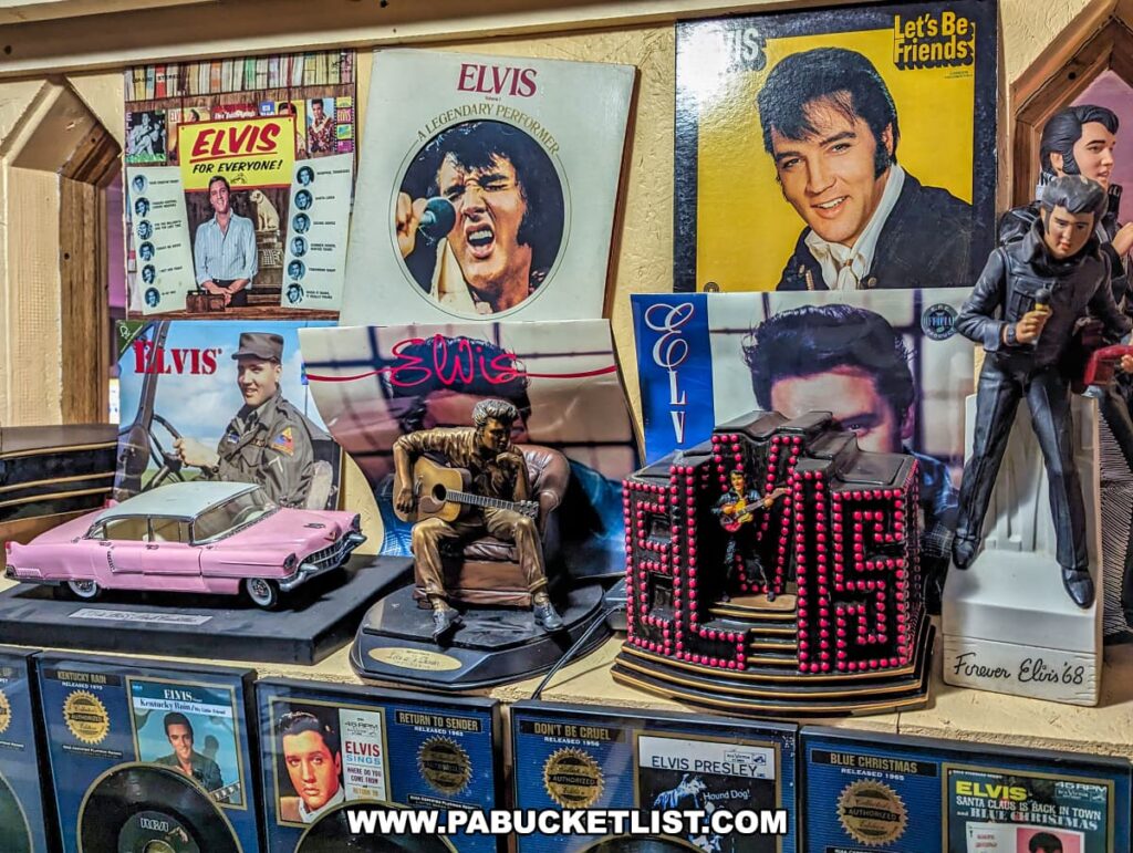 A collection of Elvis Presley memorabilia on display at Bill's Old Bike Barn museum in Bloomsburg, Pennsylvania. The exhibit includes vintage vinyl record covers showcasing different stages of Elvis's career, action figures, a pink model Cadillac, and a guitar-shaped ornament. The wall behind features images and posters of the legendary performer, creating a dedicated tribute to the King of Rock 'n' Roll within the museum's eclectic assortment of historical collectibles.