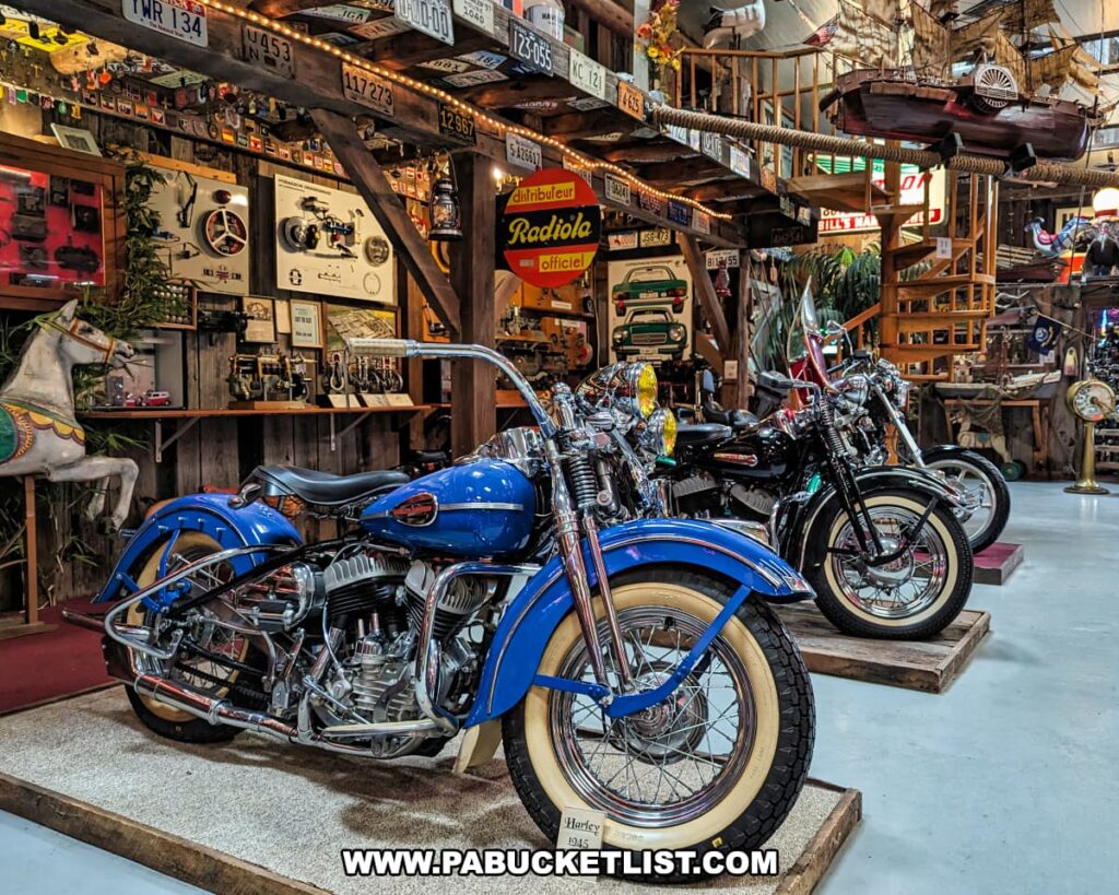 A vibrant display of classic motorcycles at Bill's Old Bike Barn museum in Bloomsburg, Pennsylvania, featuring a striking blue and white vintage motorcycle in the foreground. The collection is showcased in a rustic interior filled with antique memorabilia, including model boats, license plates, and vintage signage. The richly detailed setting creates a warm, nostalgic ambiance that celebrates the history of motorcycling.
