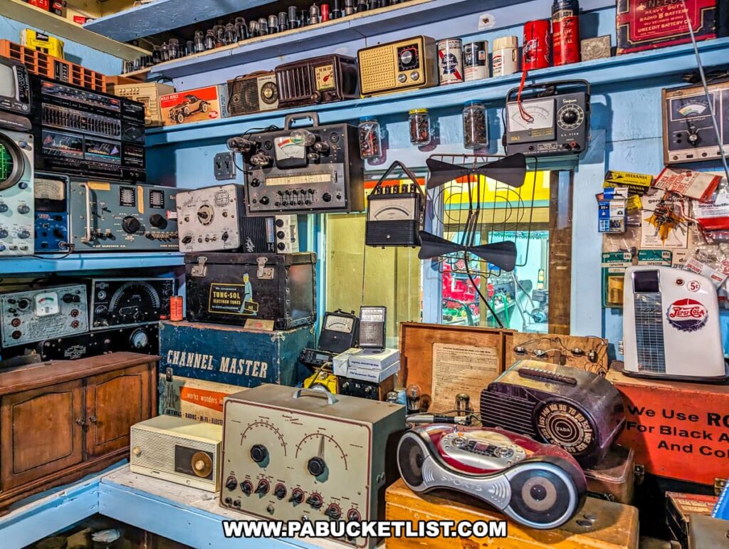 An eclectic collection of vintage electronics and memorabilia is on display at Bill's Old Bike Barn museum in Bloomsburg, Pennsylvania. The assortment includes various old radios, broadcast equipment, oscilloscopes, and signal testers arranged on wooden shelves against a blue wall. Some pieces are housed in rugged carrying cases labeled with brands like "Channel Master."