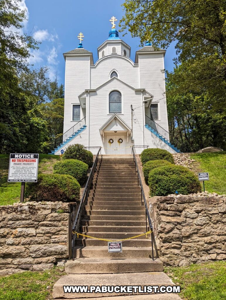 In Centralia, Pennsylvania, a striking white church with blue trim stands in sharp contrast to its solemn surroundings. The building is crowned with three gold crosses atop blue domes, a traditional feature of Ukrainian Catholic churches. A set of concrete stairs, cordoned off by yellow caution tape and flanked by 'No Trespassing' signs, leads up to the closed doors, indicating the area's isolation and abandonment. Despite the desolate backdrop of this toxic ghost town, the church remains a poignant symbol of faith and resilience amid the lingering underground coal mine fire that has ravaged the town since 1962.