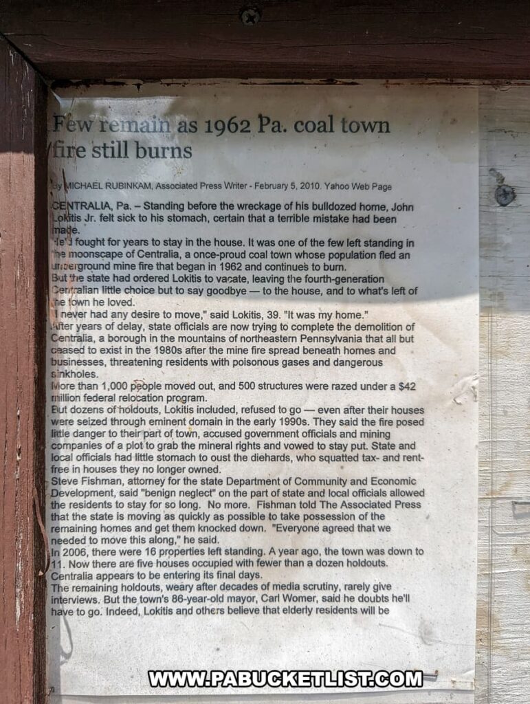 The image displays a worn newspaper article affixed to a wooden surface, discussing the lingering aftermath of the 1962 coal mine fire in Centralia, Pennsylvania. The headline reads "Few remain as 1962 Pa. coal town fire still burns," signaling the enduring presence of the fire and its effects on the town's population. The article recounts the experiences of the few residents who chose to stay despite the dangers, including a man who stood before the ruins of his bulldozed home, acknowledging the profound personal and community loss. The text goes on to detail the government's efforts to vacate the town, the demolition of homes, the state's negligence, and the steadfastness of the remaining elderly residents amidst the dwindling numbers of properties and the encroaching finality facing Centralia.