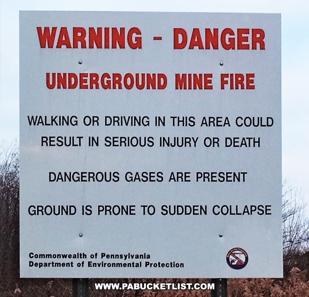 The image shows a warning sign from the Commonwealth of Pennsylvania Department of Environmental Protection, cautioning against the dangers of the underground mine fire in Centralia, Pennsylvania. It states that walking or driving in the area could result in serious injury or death due to dangerous gases and the potential for sudden ground collapse, reflecting the hazardous conditions created by the long-burning coal mine fire in this area known as a "toxic ghost town."