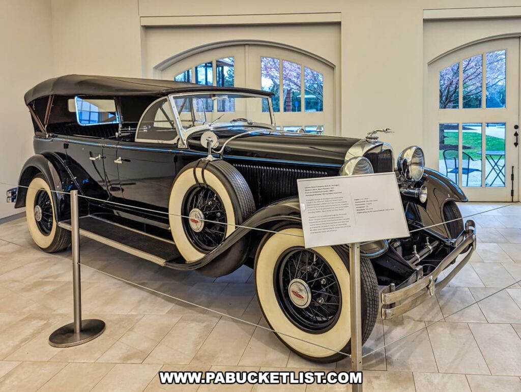 A vintage 1931 Lincoln Model K displayed inside the visitor center at the Henry Clay Frick estate in Pittsburgh, Pennsylvania. The classic car is black with cream-colored side-wall tires and features a soft convertible top, large chrome headlights, and a distinctive upright grille. It's cordoned off with stanchions and a rope, and a descriptive placard stands next to it. The vehicle rests on a tiled floor, with arched windows in the background providing a view of the outside greenery.