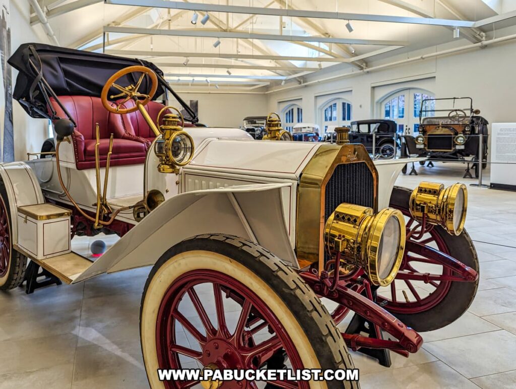 An exhibit in the Car and Carriage Museum at the Henry Clay Frick estate in Pittsburgh, Pennsylvania, featuring an antique white car with brass accents and red wheels. The car has a rich red interior and is displayed alongside other vintage automobiles in a bright, spacious gallery with a high ceiling and large windows allowing natural light.