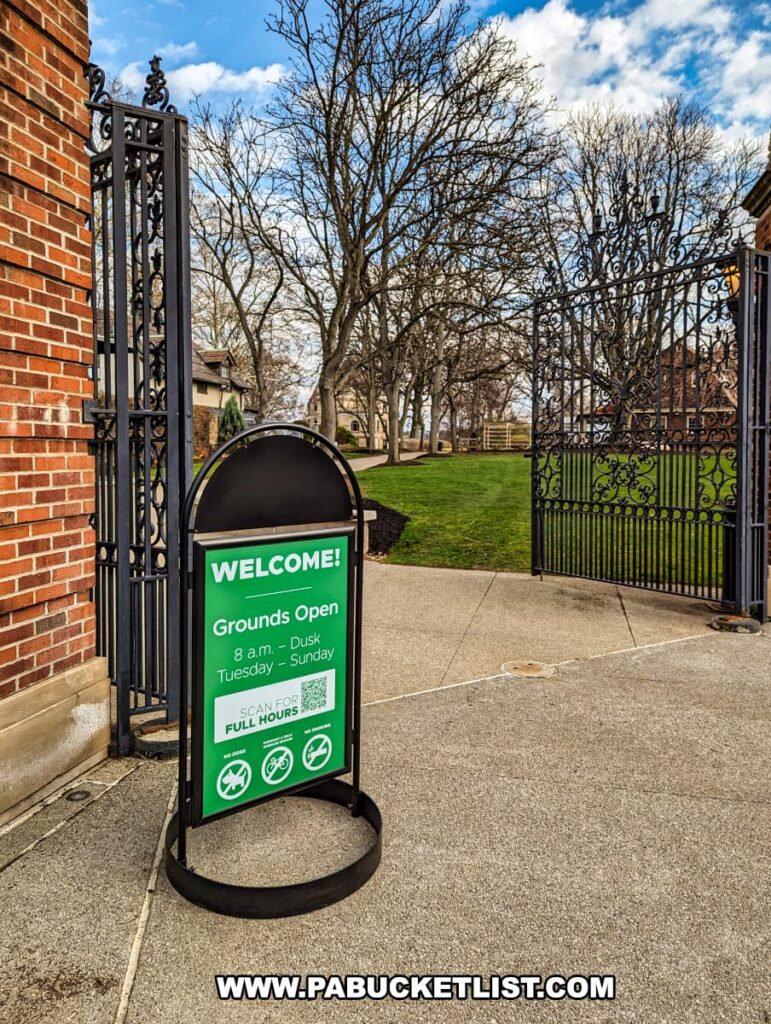 A welcoming sign at the entrance of the Henry Clay Frick estate in Pittsburgh, Pennsylvania, informs visitors that the grounds are open from 8 a.m. to dusk, Tuesday through Sunday. The sign features a green background with white lettering and icons indicating key information, including accessibility, pet policies, and photography. It stands on a paved pathway leading into the estate, flanked by leafless trees and a wrought-iron gate that exhibits intricate designs, hinting at the elegance and historical significance of the estate.