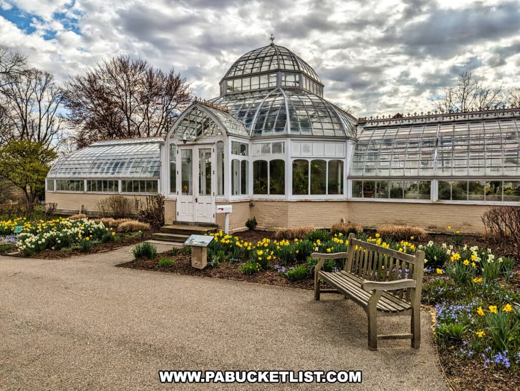 A classic greenhouse at the Henry Clay Frick estate in Pittsburgh, Pennsylvania, composed of white-framed glass panels and a central dome. It sits amidst a garden of blooming daffodils and pansies, signaling spring. In the foreground, a wooden bench invites visitors to rest and enjoy the view. The overcast sky above suggests a tranquil, early spring day at the estate.
