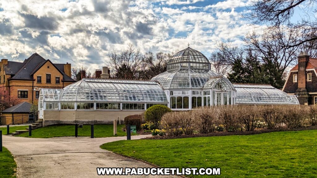 An elegant glass greenhouse at the Henry Clay Frick estate in Pittsburgh, Pennsylvania, characterized by its white framework and a central domed section. The greenhouse is surrounded by a lush green lawn and early spring blooms. Neighboring the greenhouse are traditional houses with architectural styles from the same era as the estate, creating a harmonious blend of horticultural and residential structures. The sky is partly cloudy, suggesting a cool spring day, ideal for a leisurely visit to the historical grounds.