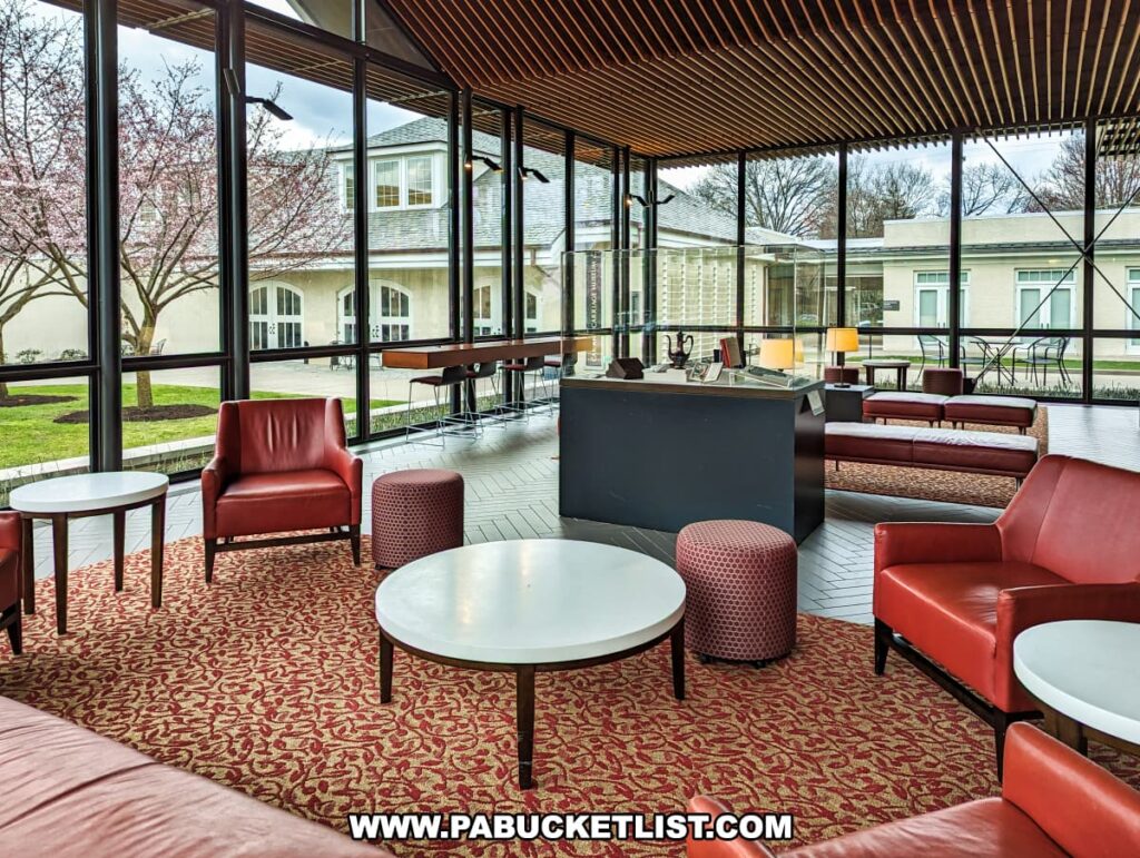 This image features the interior of the visitor center at the Henry Clay Frick estate in Pittsburgh, Pennsylvania. The space is filled with natural light streaming in through the large floor-to-ceiling windows, providing a view of the blooming cherry trees outside. The room is furnished with elegant, modern furniture, including red leather armchairs, white round tables, and red cylindrical stools, all set upon a carpet with a rich red and beige pattern. The design also includes a sleek black countertop area, possibly a reception or a bar, against the backdrop of the warm wooden slat ceiling and matching vertical blinds, creating a welcoming space for visitors to relax and gather.