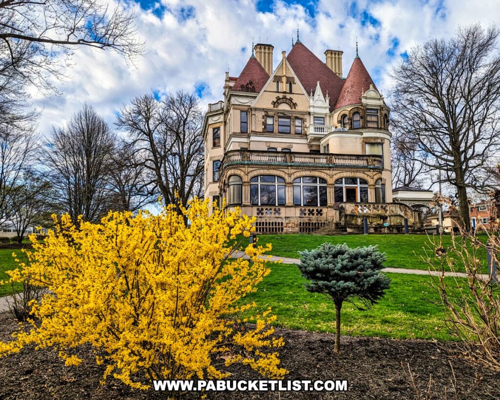 The Henry Clay Frick estate in Pittsburgh, Pennsylvania, during spring, showcasing the stately Clayton mansion, a Victorian-era chateau with decorative gables, prominent chimneys, and a wrap-around porch. The mansion is set against a backdrop of leafless trees and a cloudy sky. In the foreground, vibrant yellow forsythia blooms add a pop of color to the scene, while a small conifer provides greenery to the well-kept garden.
