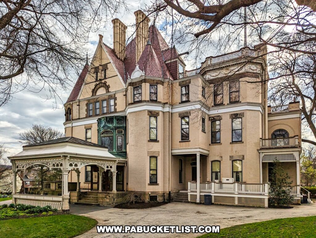 The Frick estate mansion in Pittsburgh, Pennsylvania, stands majestically with its elaborate Victorian architecture, featuring steep gables, decorative trim, and an ornate enclosed porch. The creamy beige façade is complemented by the brownstone accents and slate roof, while the surrounding landscape is beginning to show signs of spring with leafless trees and a bright green lawn.