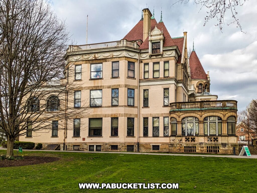 The Clayton mansion at the Henry Clay Frick estate in Pittsburgh, Pennsylvania, showcases its elegant Victorian architecture with a cream-colored façade, distinctive red roof peaks, and a rounded, glass-paned enclosed porch. The historical beauty of the building is enhanced by its stately windows and decorative stone trim. The mansion is set against a backdrop of bare trees and a gray sky, with a well-kept lawn in the foreground, inviting visitors to explore its grandeur and history.