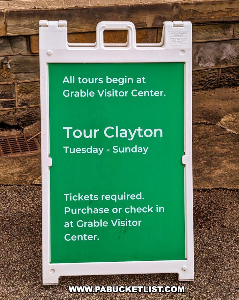 A green and white informational sign at the Henry Clay Frick estate in Pittsburgh, Pennsylvania, indicates that all tours begin at the Grable Visitor Center. It notes that the Tour Clayton is available from Tuesday to Sunday and that tickets are required, which can be purchased or checked in at the Grable Visitor Center. The sign is placed on a sidewalk against a stone wall, providing clear directions for visitors.