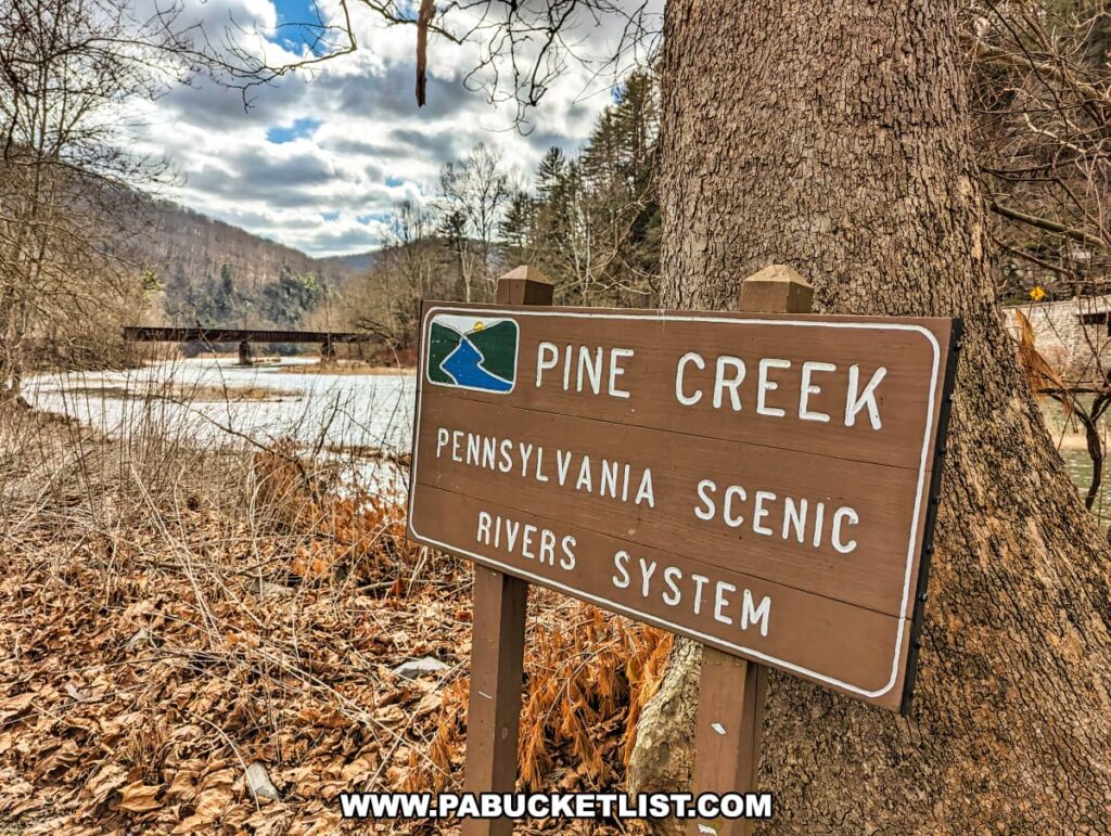 A wooden sign reading 'Pine Creek Pennsylvania Scenic Rivers System' positioned against a tree with a scenic backdrop of Pine Creek and a covered bridge in Tioga County, Pennsylvania. The creek appears calm and reflects the surrounding landscape. Leafless trees and dried leaves on the ground suggest late fall or winter. The image captures the tranquil essence of the area near the Mid State Trail, with a partly cloudy sky above.