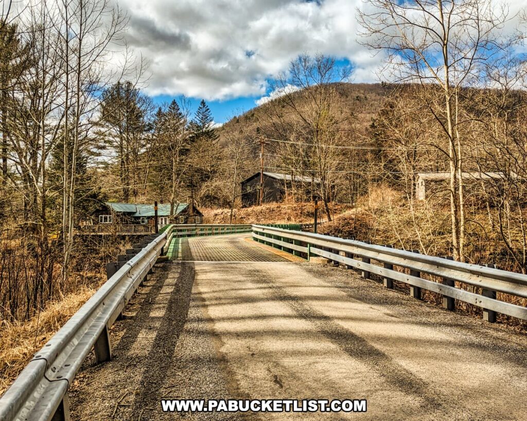 A view of a rural open grate bridge in Blackwell, Tioga County, Pennsylvania, leading towards a collection of cabins nestled among leafless trees. The metal bridge features a sturdy guardrail and a patterned surface allowing views of the creek below. The landscape suggests early spring or late fall, with a hilly backdrop, a mix of evergreen and bare deciduous trees, and a partly cloudy sky. Shadows from the bridge's railings stripe the road, adding texture to the scene near the Mid State Trail.