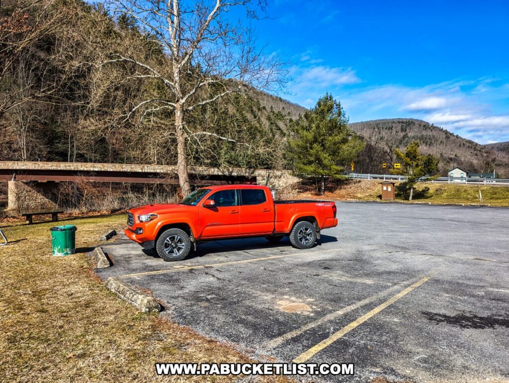 A bright orange Toyota Tacoma pickup truck is parked in a deserted asphalt parking lot near Gillespie Point in Tioga County, Pennsylvania. Leafless trees and evergreens border the area, with a bridge crossing Pine Creek visible in the background. The landscape is indicative of late fall or early spring, with a clear blue sky overhead and a backdrop of gentle hills in the distance, suggesting the peaceful seclusion of the area along the Mid State Trail.