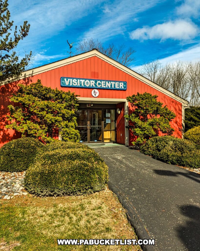 The Visitor Center at Montour Preserve near Danville, Pennsylvania, a welcoming red building with white trim and a sign that reads "VISITOR CENTER." It is a sunny day with blue skies, and the center is surrounded by well-maintained shrubbery and trees. The asphalt pathway leads to the front door, inviting visitors to explore the educational resources and exhibits housed within.