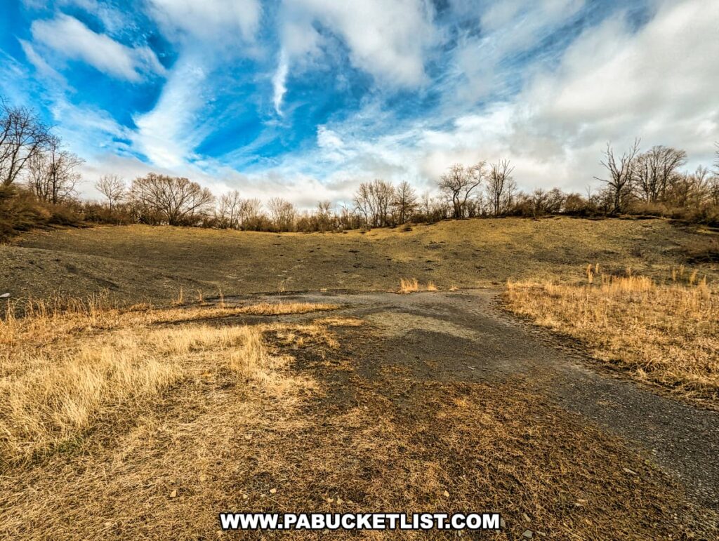 The landscape of the Montour Preserve fossil pit near Danville, Pennsylvania, featuring a wide, barren field of grayish sediment with a curved path leading through it. Sparse vegetation and leafless trees surround the area, typical of late fall or early spring. Overhead, the sky is dramatic with swirling white clouds against a bright blue backdrop, adding a dynamic contrast to the earthy tones below.