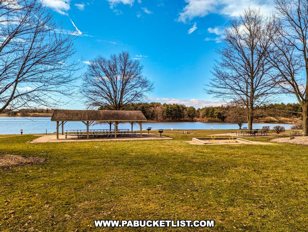 A lakeside picnic area at Montour Preserve near Danville, Pennsylvania, with a covered pavilion housing multiple picnic tables. The pavilion is positioned on a concrete pad overlooking the lake, providing a scenic spot for gatherings. Barren trees suggest the photo was taken in late fall or early spring. A vibrant green lawn stretches out in front of the pavilion, leading to the lake's edge.The sky is a bright blue with scattered clouds, indicating a clear day.
