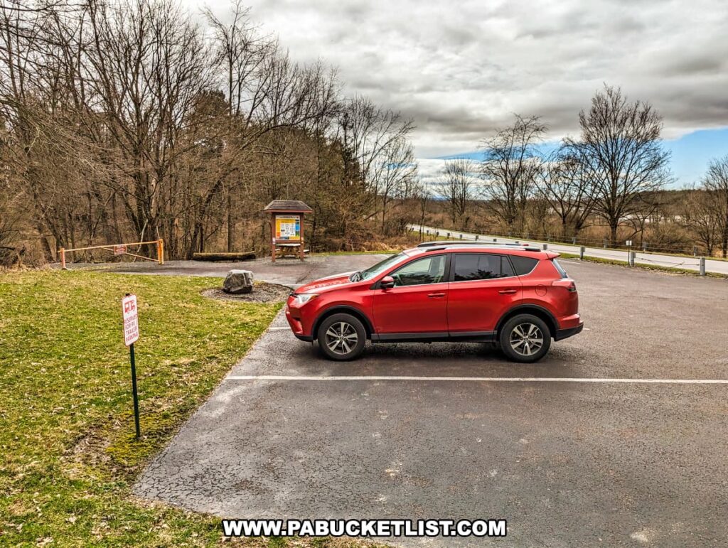 A red SUV parked in a paved parking area near the Montour Preserve fossil pit close to Danville, Pennsylvania. The parking lot is marked by white lines with a "Parking By Permit Only" sign visible in the foreground. In the background, a wooden gate and information kiosk can be seen, marking the start of a trail, likely leading to the fossil pit. The trees are barren, suggesting the photo was taken in late fall or winter. The sky is overcast, giving the setting a moody atmosphere.