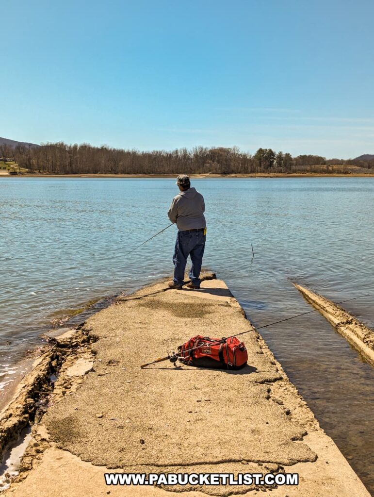 A person stands fishing at the end of the Sunken Highway at Bald Eagle State Park, with a fishing rod in hand and a tackle bag resting on the concrete path. The old stretch of US Route 220 extends into Sayers Lake, surrounded by calm waters and a peaceful landscape under a clear blue sky. The road, showing signs of decay and resilience, offers a unique fishing spot, blending history with leisure against the scenic backdrop of the park.