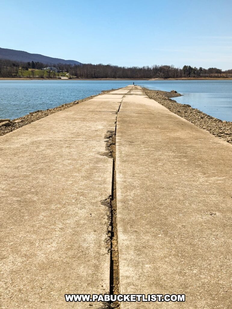 A long, narrow concrete roadway, bordered by rocky edges, extends directly into the horizon on Sayers Lake at Bald Eagle State Park. The clear blue sky above and the calm waters to either side frame the Sunken Highway, which once carried travelers along Old US Route 220. Now, it lies as a silent witness to the past, leading towards the distant township of Howard, Pennsylvania, with a solitary fisherman in the distance enjoying the view.