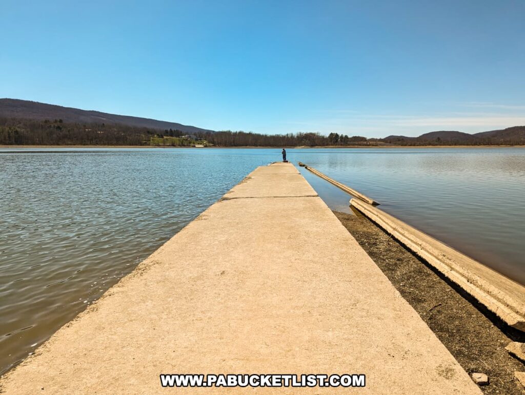 The concrete remains of the Sunken Highway extend into the tranquil waters of Sayers Lake at Bald Eagle State Park, providing a striking linear perspective against the natural surroundings. A single figure stands in the distance on the road, framed by the expansive blue sky and the serene lake, with the gentle rolling hills and the outline of a community near Howard, PA, providing a picturesque backdrop.