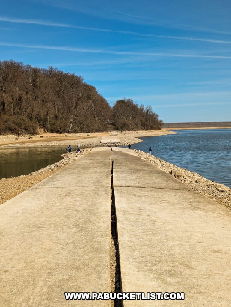 The image showcases the historic Sunken Highway at Bald Eagle State Park near Howard, PA, a straight concrete road that cuts across Sayers Lake. Visitors can be seen on either side of this once-submerged stretch of Old US Route 220, enjoying the unique landscape where the road divides the water, leading the eye toward the distant tree-lined shore and blue sky above.