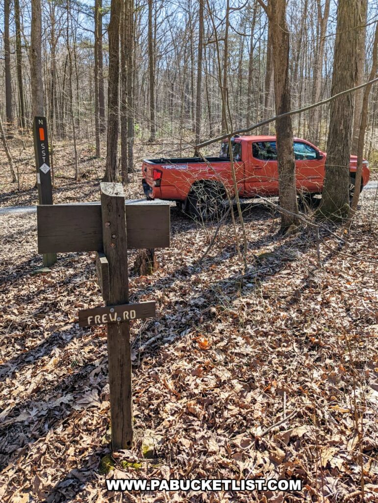 A red pickup truck parked beside the forested trailhead at Frew Road, which leads to the Three Sisters Rock Formation on the Standing Stone Trail in Huntingdon County, Pennsylvania, with trail markers visible on a tree and a wooden sign indicating "FREW RD." in the foreground.