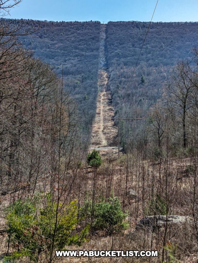 A clear-cut path through a forested landscape for power lines, leading up a hill, near the Three Sisters Rock Formation along the Standing Stone Trail in Huntingdon County, Pennsylvania, with early spring vegetation and a blue sky above.