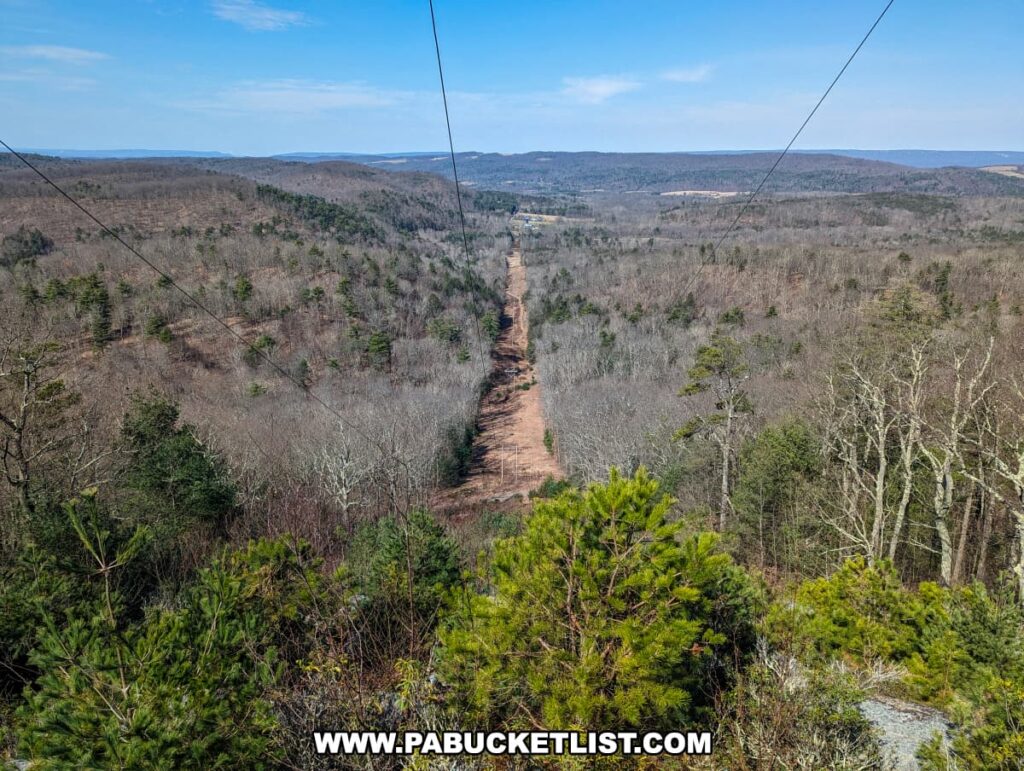 A vista of a cleared path for power lines running through a dense woodland area, viewed from the Three Sisters Rock Formation along the Standing Stone Trail in Huntingdon County, Pennsylvania. The path cuts a straight line through the forest, contrasting with the natural undulating terrain of the surrounding hills under a wide blue sky.